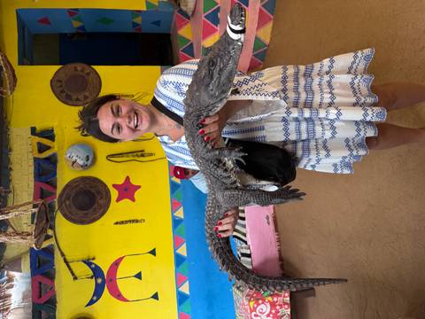 A smiling woman in a patterned dress holds a small crocodile inside a brightly painted Nubian home.