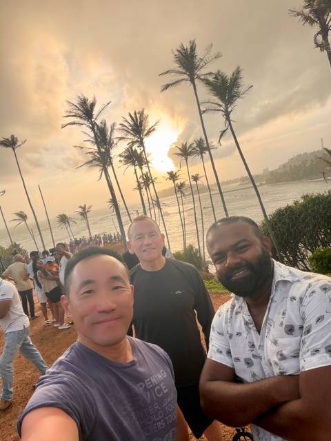 Selfie of three travelers at a palm-lined coastal headland during golden sunset with crowd behind