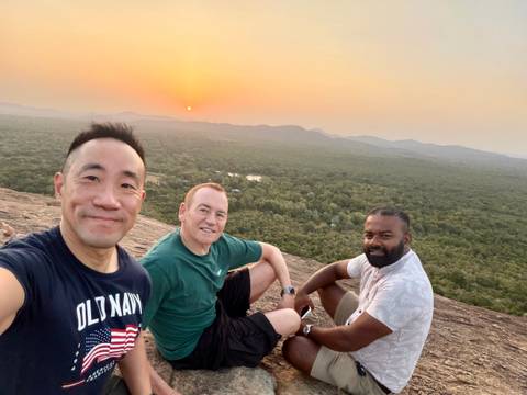 Three travelers resting on a rocky summit overlooking forested plain at sunset