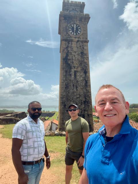 Three travellers pose for a selfie beside a historic stone tower overlooking a coastal landscape.