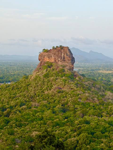 Iconic Sigiriya Rock fortress rising above dense Sri Lankan jungle with distant hazy hills.
