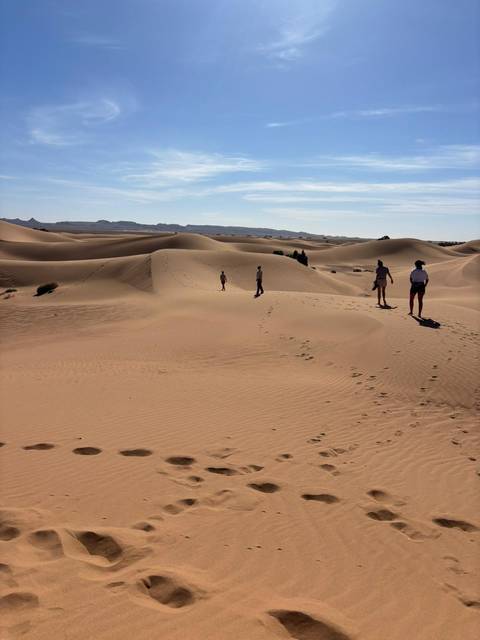 Travelers walking atop rolling sand dunes with footprints visible across the desert.