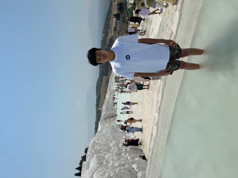 Teen boy posing barefoot in the shallow turquoise pools of Pamukkale with many tourists walking along the white calcium terraces in the background.