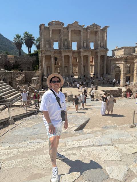 Smiling man in hat and sunglasses stands in front of the ancient Library of Celsus ruins at Ephesus with crowds exploring the archaeological site.