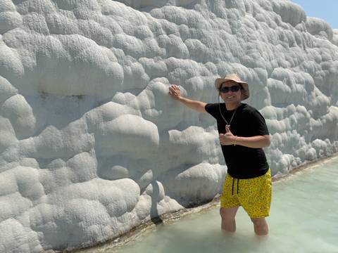 Man in yellow swim shorts touches the rippled white calcium wall of Pamukkale while standing in shallow water.