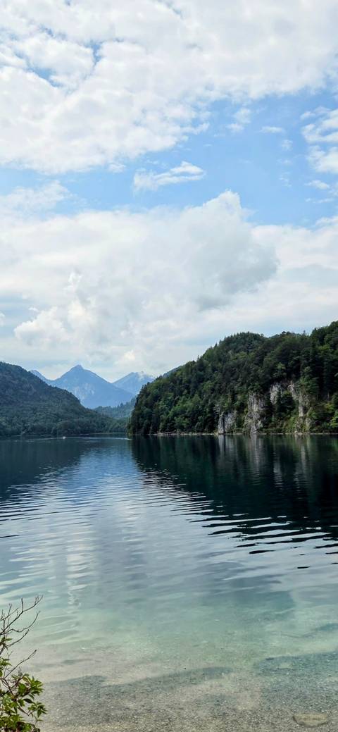 Quiet alpine lake reflects tree-covered hills and distant peaks beneath a cloudy Bavarian sky.