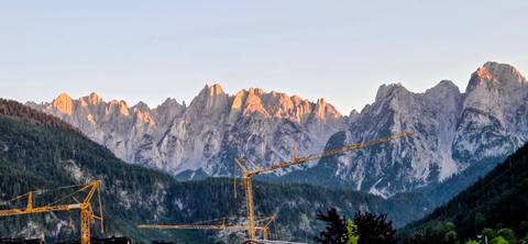 Construction cranes stand before rugged alpine peaks glowing in the warm light of sunset.