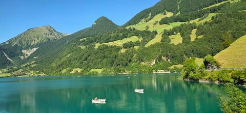 Rowboats drift on a crystal-clear turquoise alpine lake bordered by rolling green hills and forests.