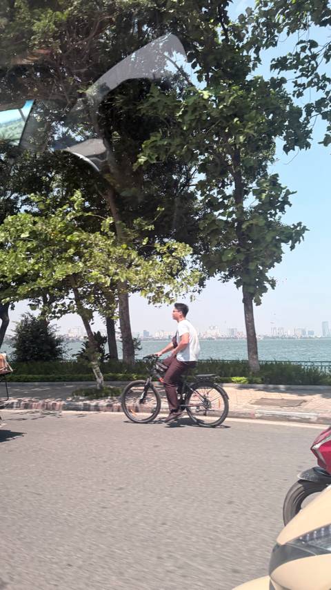 Man cycling along lakeside path with city skyline across the water, motion blur in foreground trees.