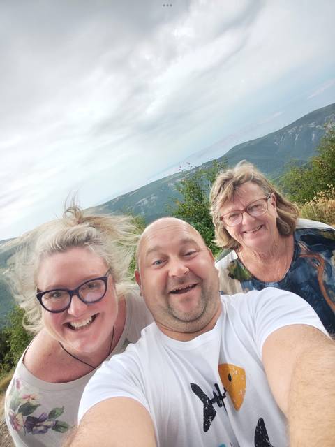 Close selfie of three happy travelers with windy mountain scenery in the background.