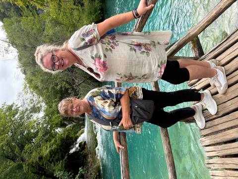 Two women stand on a wooden bridge over turquoise water with waterfalls and forest behind.