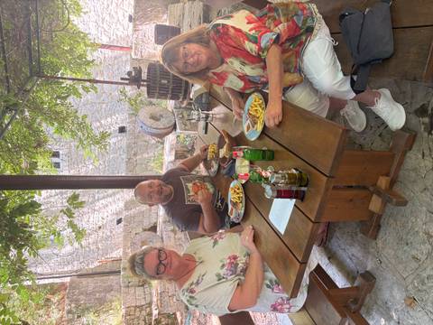 Three travelers enjoy beer and fries at a rustic outdoor wooden table under leafy shade.