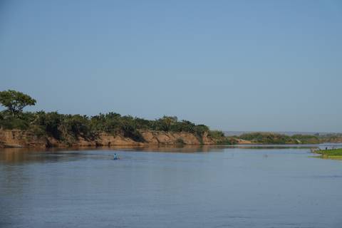 Wide calm river bordered by earthy banks and trees with a lone paddler gliding across the water under clear blue sky.