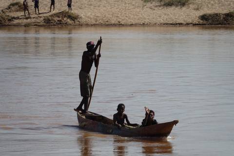 Traditional wooden canoe with three locals, one standing to pole, crossing a brown river.