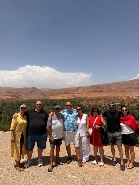 Travellers pose on a terrace overlooking the lush oasis and earthen buildings of a Moroccan valley with arid mountains beyond.