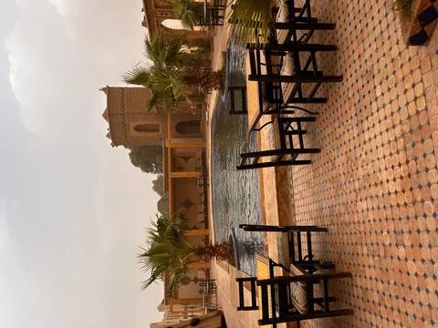 Courtyard pool flanked by palm trees and a mud-brick tower, with woven chairs on tiled terrace.