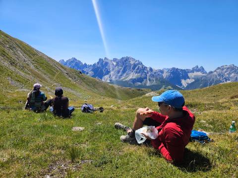 Hikers rest on an alpine meadow enjoying lunch while admiring jagged Dolomite peaks under a clear sky.
