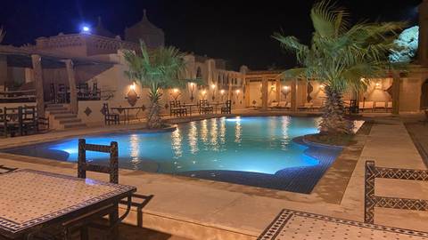 An atmospheric riad courtyard at night with a lit turquoise swimming pool and palm trees.