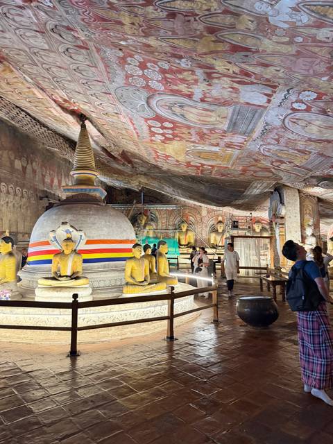 Ornate cave temple interior with golden Buddha statues and vivid painted ceiling, visitors exploring the hall