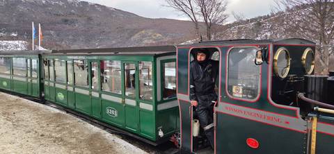 Vintage green train with a bundled conductor standing in the open doorway against a snowy mountain backdrop.