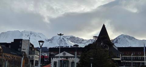 Snow-capped mountains towering over a small coastal city under a moody cloudy sky.