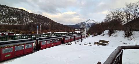 Historic red train carriages steaming through a snowy forested valley with peaks in the distance.