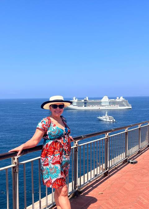 Woman on cliff-side railing with cruise ship and yacht on deep blue sea below