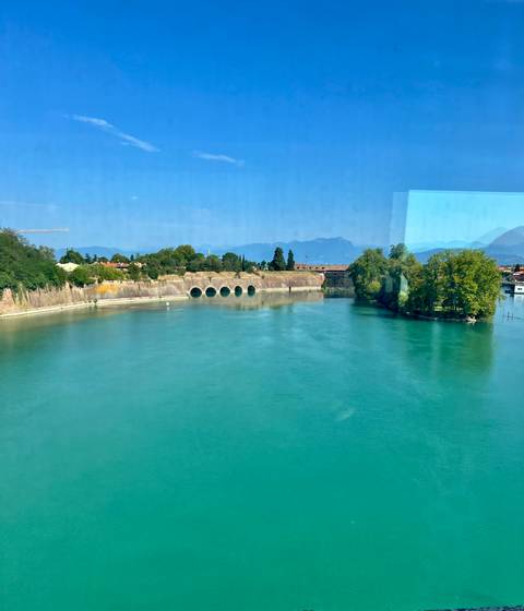 Calm turquoise river bending past fortified walls with distant mountains under blue sky