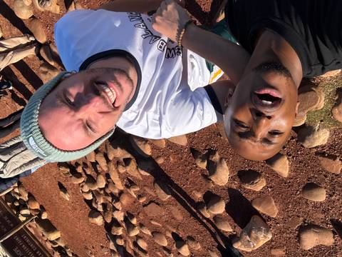 Two young men laugh while lying on reddish ground strewn with stones, taking a playful close-up selfie.