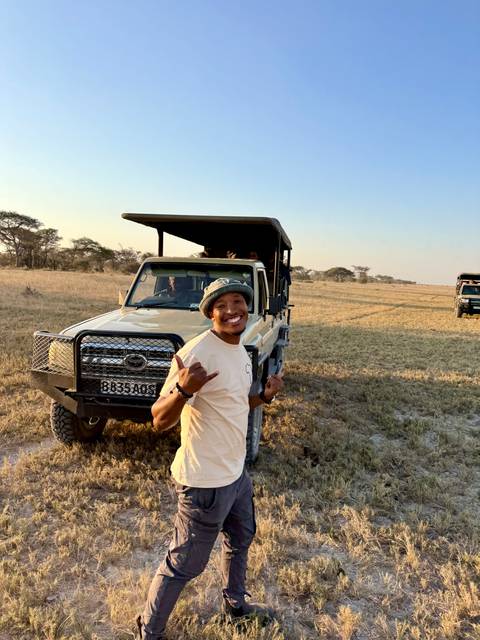 A safari guide stands smiling beside an open 4x4 in a dry grassland as the evening light falls.