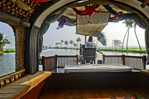 View from inside a traditional Kerala houseboat looking out over calm backwaters lined with palm trees as the captain steers.