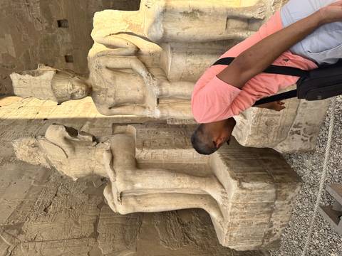 Visitor bending to study colossal seated statues carved with hieroglyphs inside an ancient Egyptian temple.