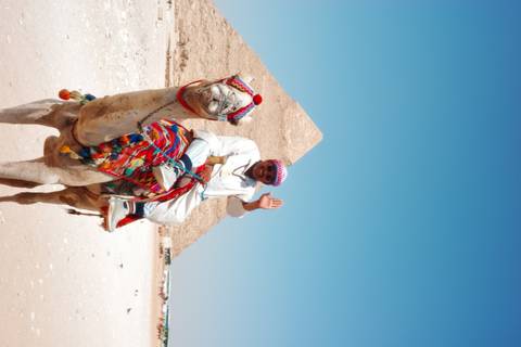 Smiling man in traditional attire riding a decorated camel with a pyramid rising in the background under clear skies.