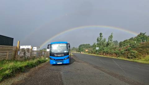 Blue tour bus parked beside a road beneath a full rainbow and dark sky in rural Scotland