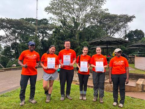 Hikers wearing matching orange shirts proudly display Kilimanjaro certificates at a lush park viewpoint