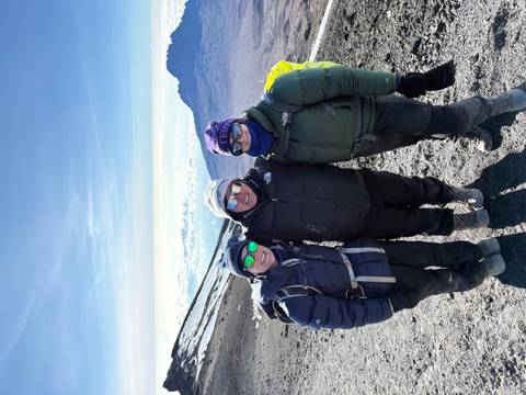 Three climbers wearing goggles and heavy jackets standing on volcanic scree with distant crater and sky