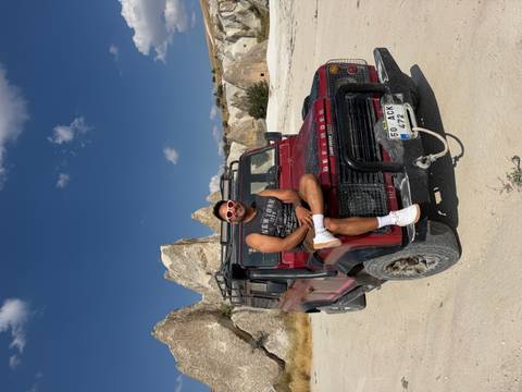 Young man in sunglasses posing on a red 4x4 jeep with Cappadocia rock formations behind