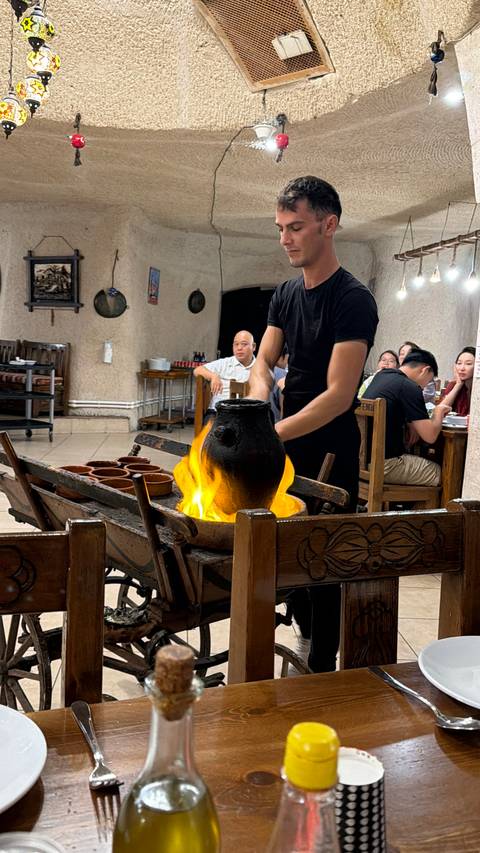 Restaurant scene where a waiter flames a clay pot over fire while diners watch