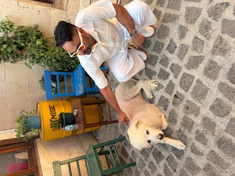 Man in white clothes petting a light-colored dog on a stone courtyard with rustic chairs