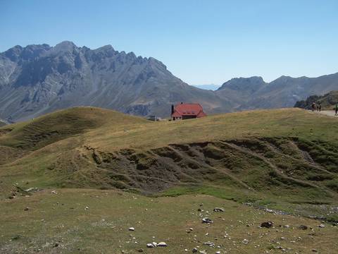 Alpine meadow with a lone red-roofed hut set against jagged grey peaks.