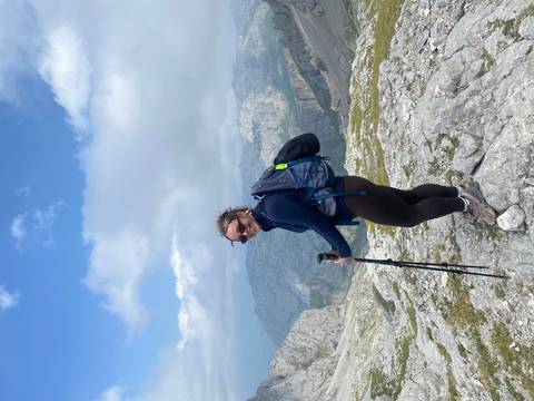 Female hiker with trekking poles standing on a rocky summit above cloud-draped mountains.