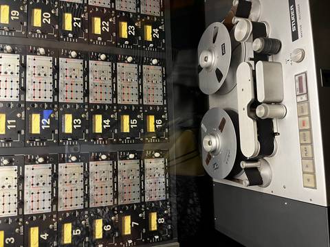 Close-up of a vintage reel-to-reel tape recorder displayed in front of a wall of analog audio equipment meters and dials.