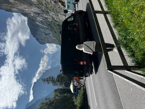 A mountain valley road bordered by dramatic cliffs and peaks; a black minibus passes while an older woman admires the alpine scenery.
