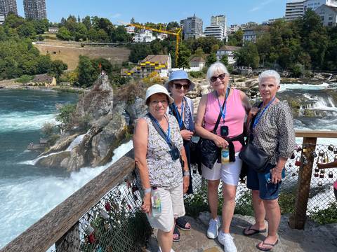 Four smiling women pose on a wooden viewing deck overlooking the powerful Rhine Falls cascading around a rocky islet.