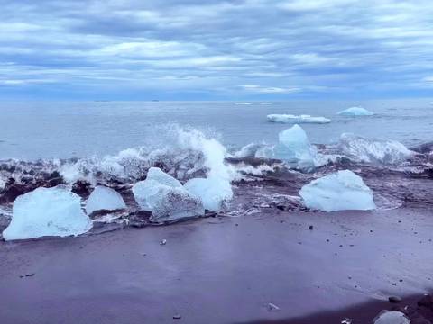 Waves crash over luminous blue ice chunks scattered on a black-sand beach under cloudy skies.