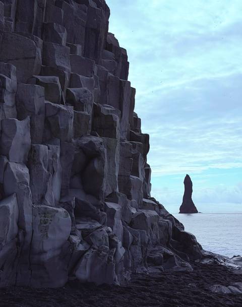 Tall basalt sea stack rises offshore beside dramatic hexagonal cliff columns under blue sky.