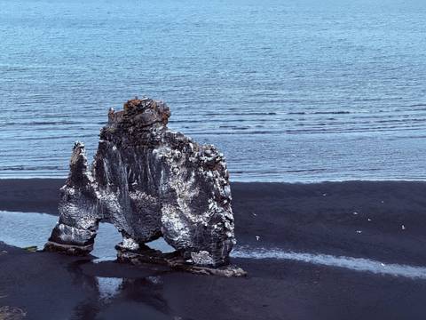 Rocky sea arch Hvitserkur stands against black-sand shoreline and rippled blue water.