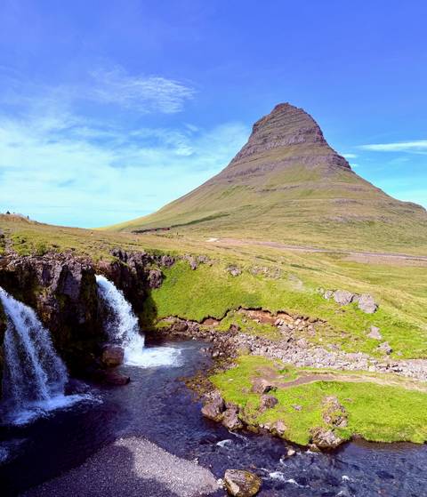Kirkjufell mountain rises above a cascading waterfall under a bright blue sky.