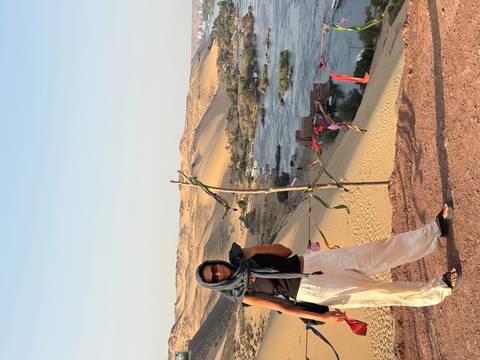 Woman with scarf stands on sandy ridge above lush Nile riverbank and Nubian village.