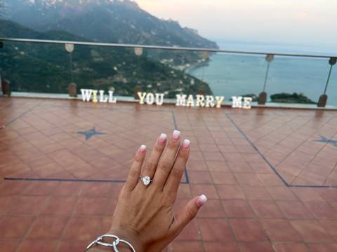 Hand with engagement ring held up against a coastal panorama with marquee letters spelling 'WILL YOU MARRY ME'.